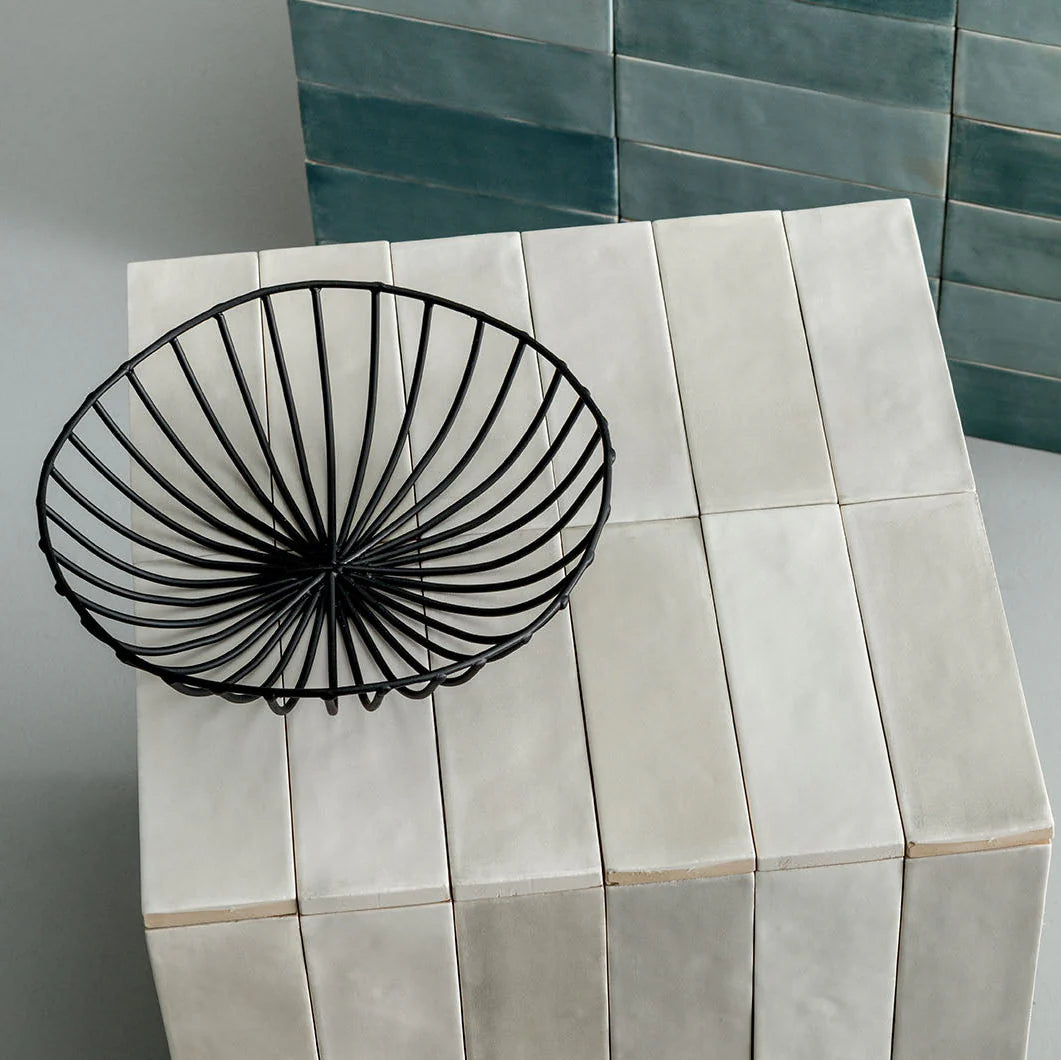 Tint Dune Matt Subway 52x160mm white ceramic tiles covering a cube pedestal styled with a black wire bowl against teal zellige tiles.