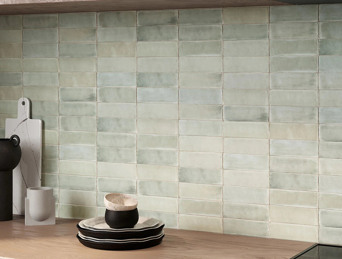 Tint Sea Glass Matt Subway 52x160mm tiles installed as a full kitchen splashback in soft sage green, styled with stacked ceramic plates and a marble chopping board.
