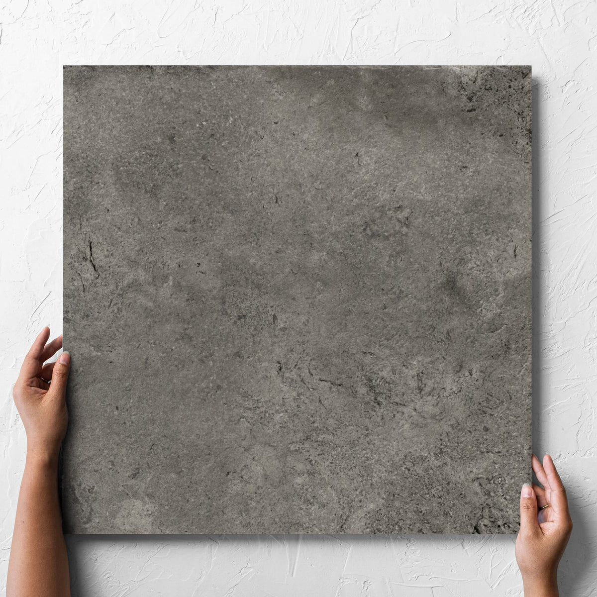 Hand holding a single Toulouse Dark Grey Amber 600x600mm tile against a white rendered wall, showing the matt surface