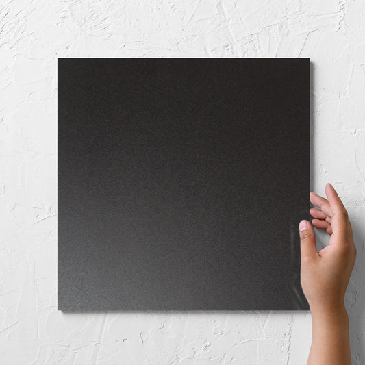 Hand holding a single Unicolour Black Matt 300x300mm tile against a white rendered wall, showing the black matt surface