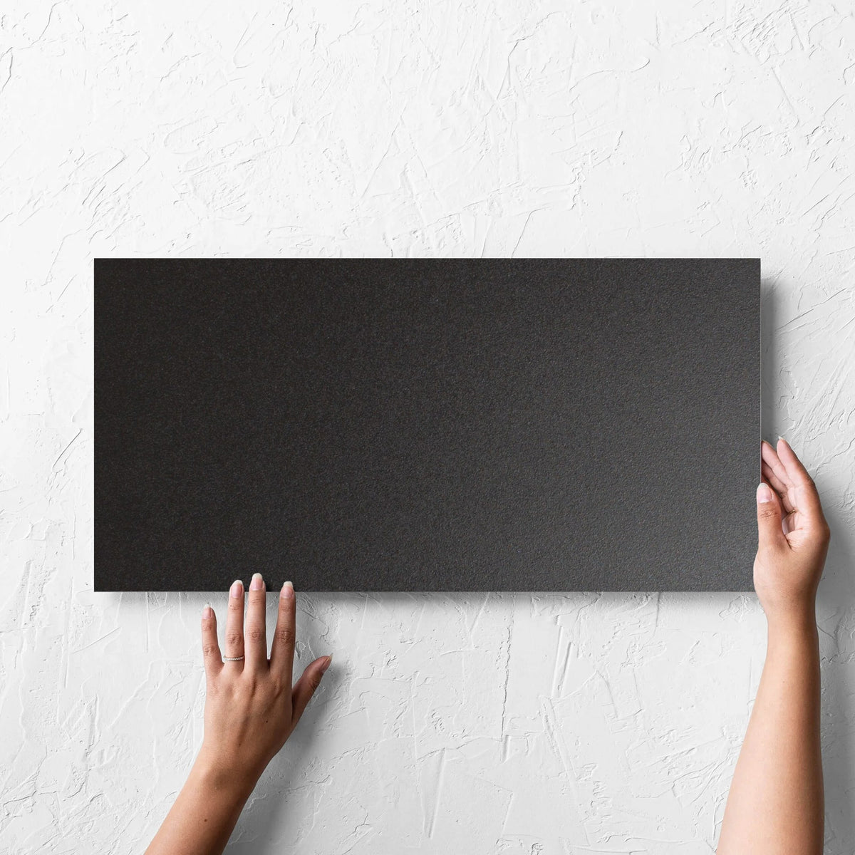 Hand holding a single Unicolour Black Matt 300x600mm tile against a white rendered wall, showing the black matt surface
