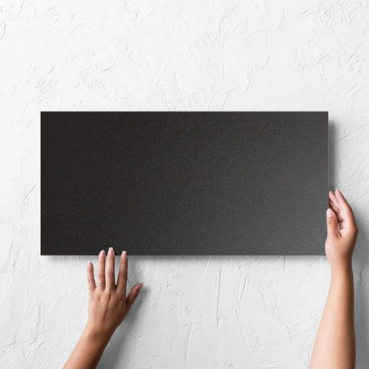 Hand holding a single Unicolour Black Matt 300x600mm tile against a white rendered wall, showing the black matt surface