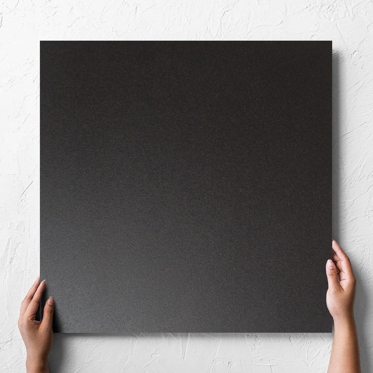 Hand holding a single Unicolour Black Matt 600x600mm tile against a white rendered wall, showing the black matt surface