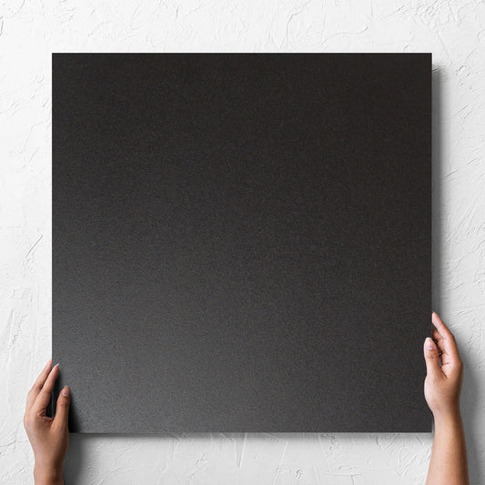 Hand holding a single Unicolour Black Matt 600x600mm tile against a white rendered wall, showing the black matt surface