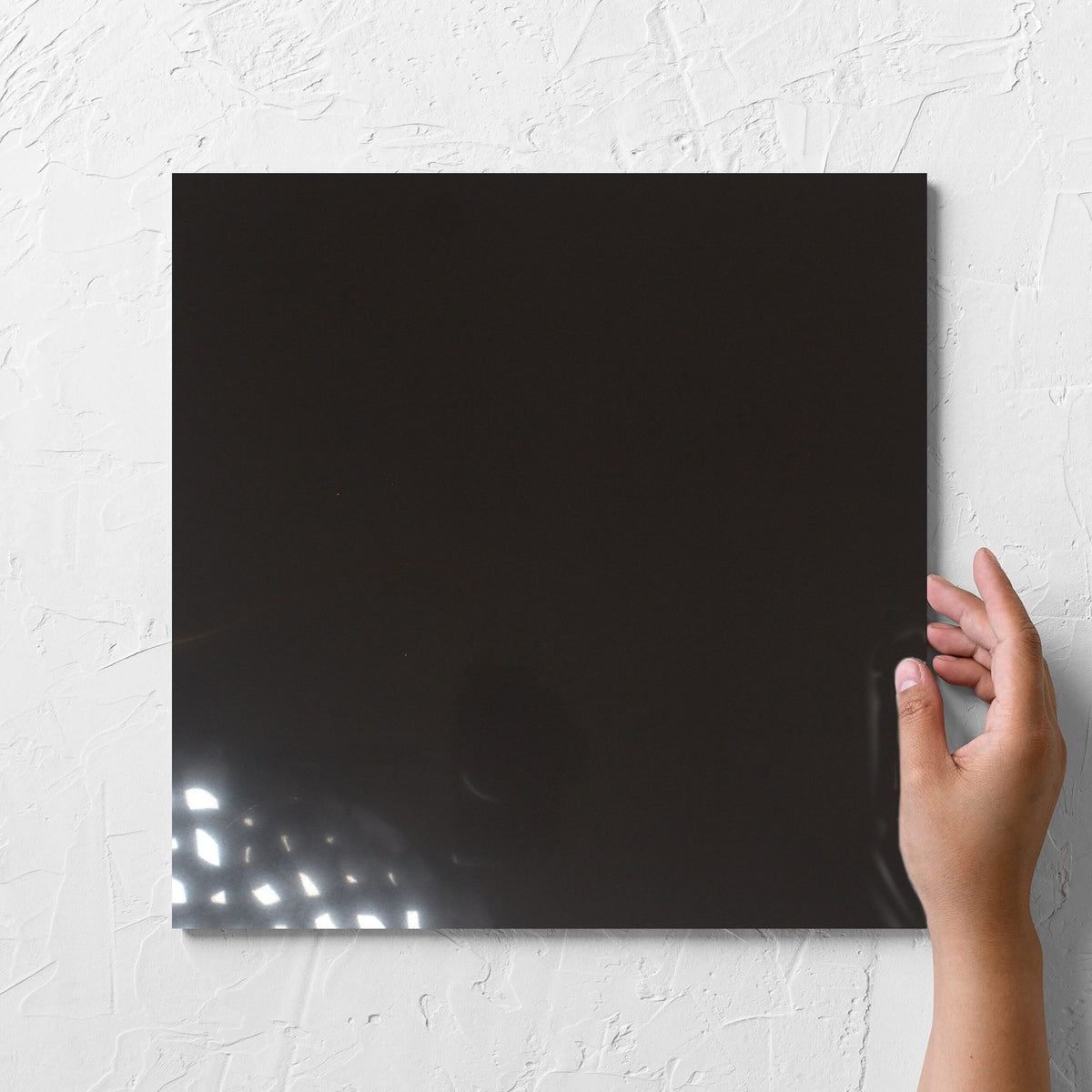 Hand holding a single Unicolour Black Polished 300x300mm tile against a white rendered wall, showing the black polished surface