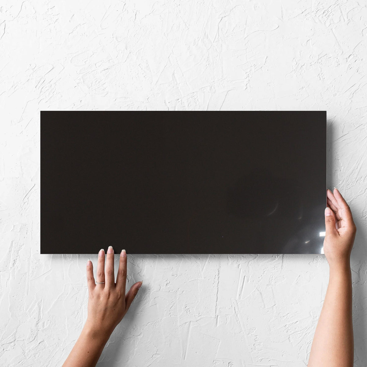 Hand holding a single Unicolour Black Polished 300x600mm tile against a white rendered wall, showing the black polished surface