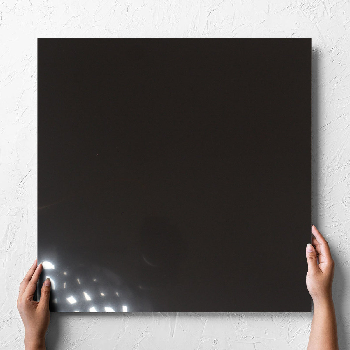 Hand holding a single Unicolour Black Polished 600x600mm tile against a white rendered wall, showing the black polished surface