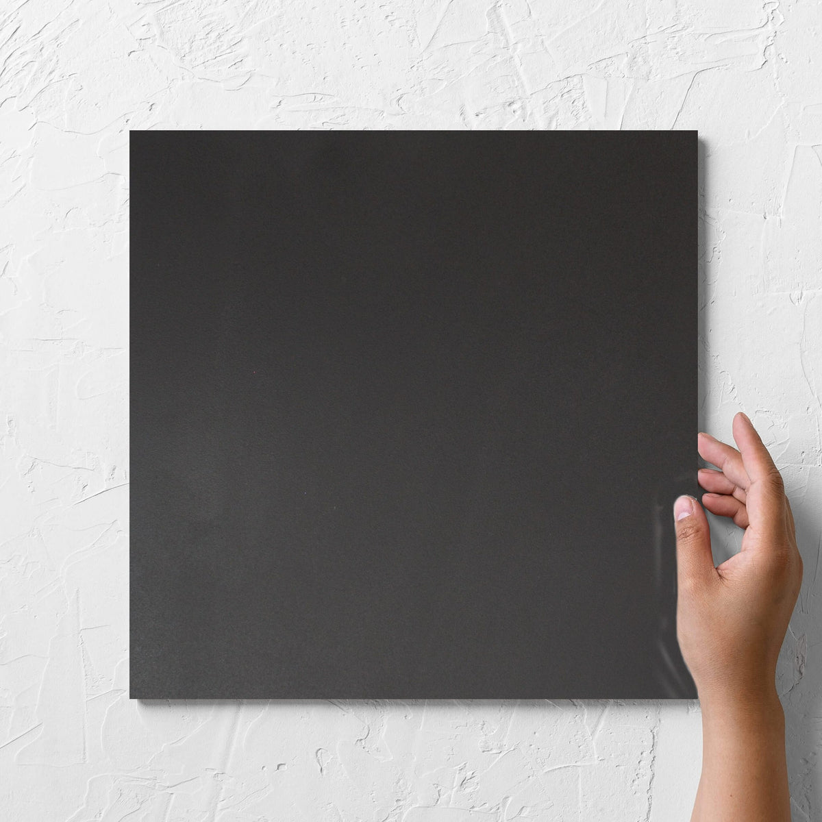 Hand holding a single Unicolour Black R11 300x300mm tile against a white rendered wall, showing the black r11 surface
