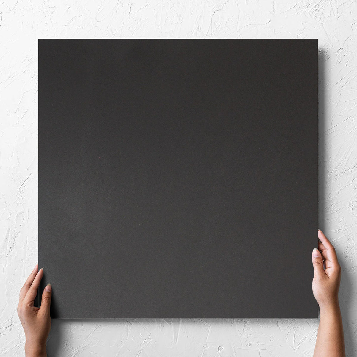 Hand holding a single Unicolour Black R11 600x600mm tile against a white rendered wall, showing the black r11 surface