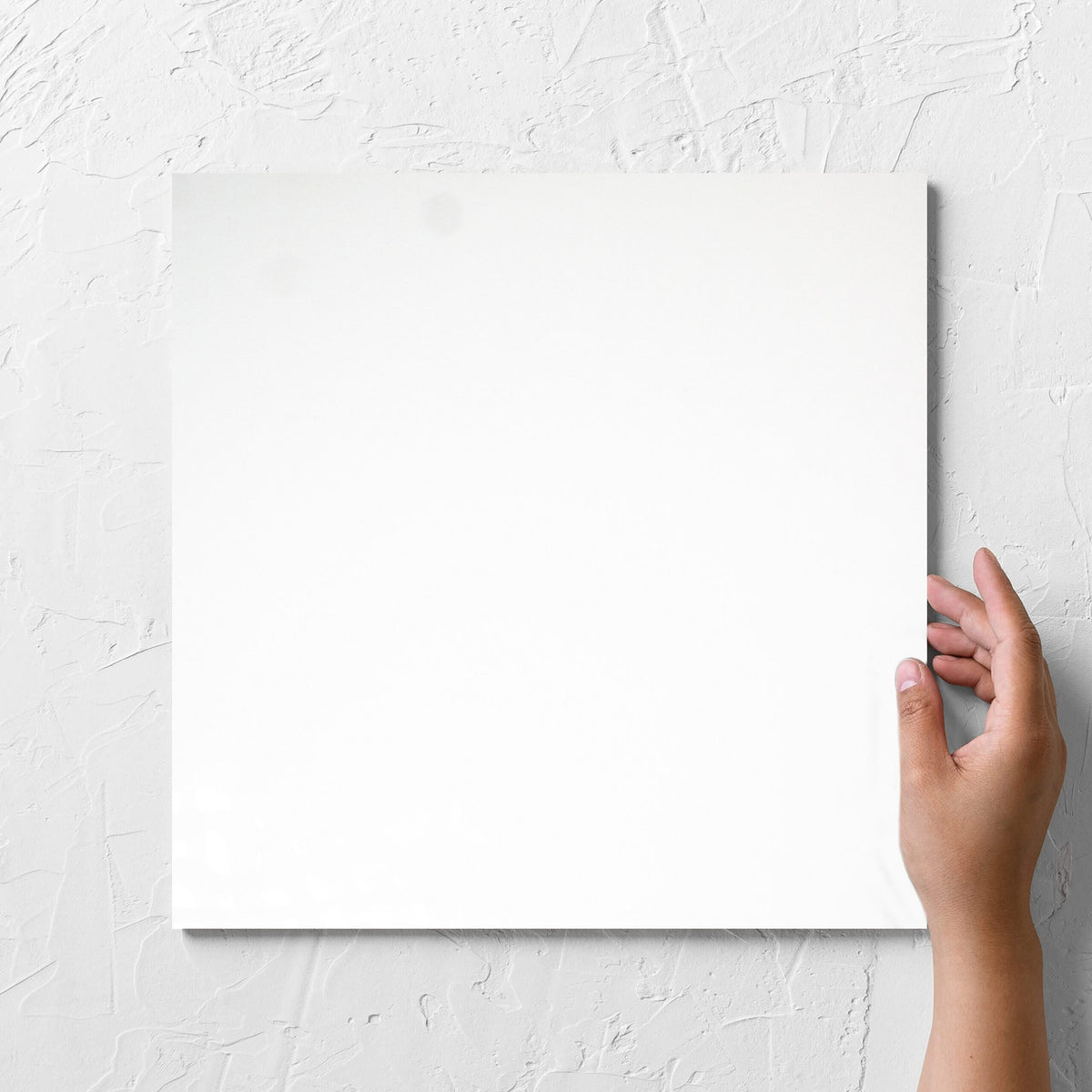 Hand holding a single Unicolour White Polished 300x300mm tile against a white rendered wall, showing the white polished surface