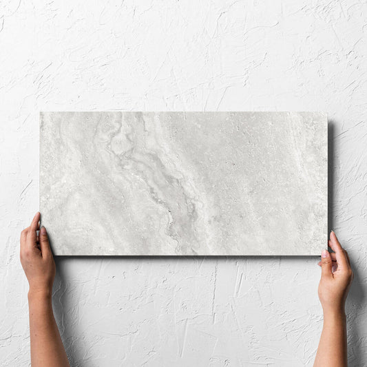 Hand holding a single Vatican Silver Cross Cut Travertine 300x600mm tile against a white rendered wall, showing the matt surface