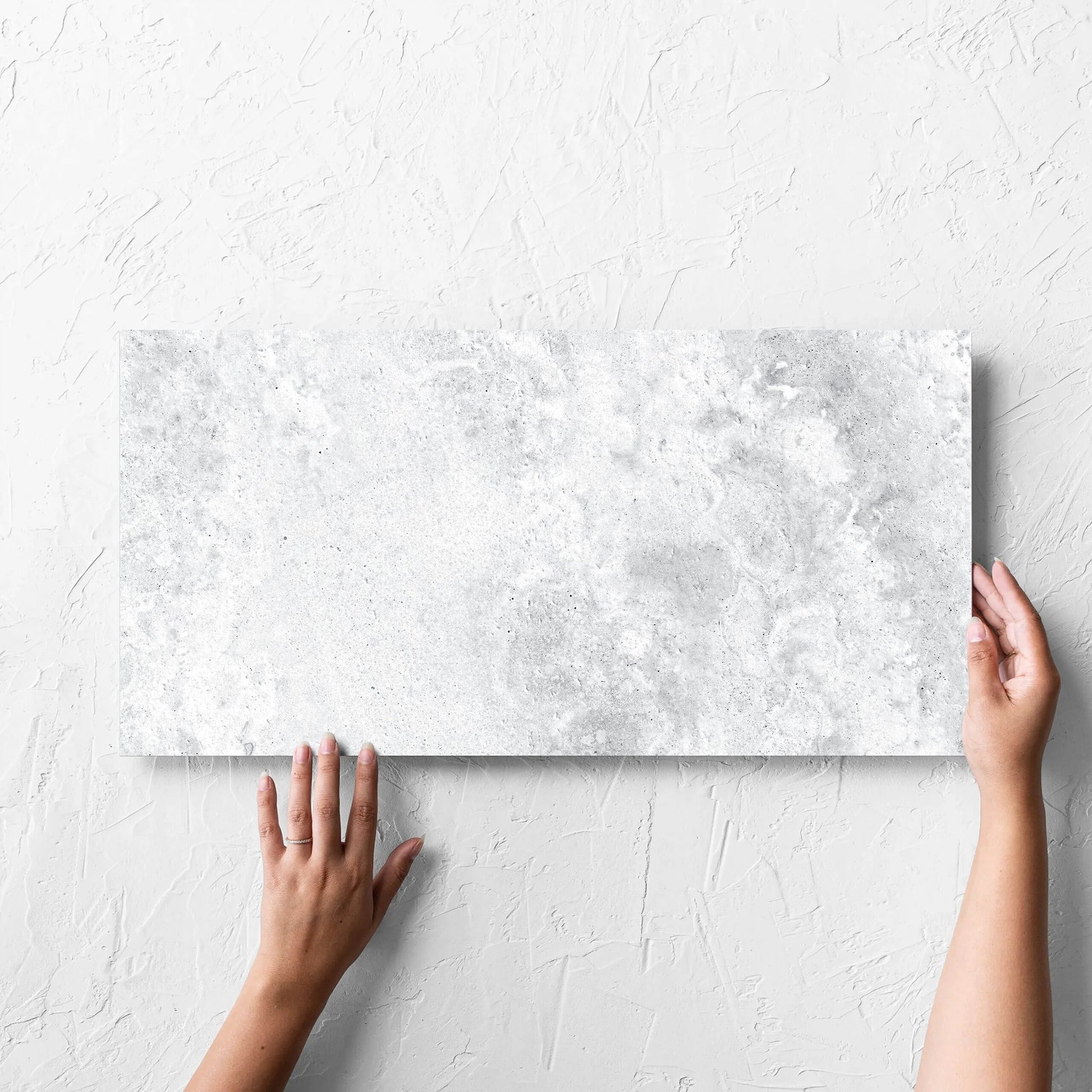 Hand holding a single Vatican White Cross Cut Travertine 300x600mm tile against a white rendered wall, showing the matt surface