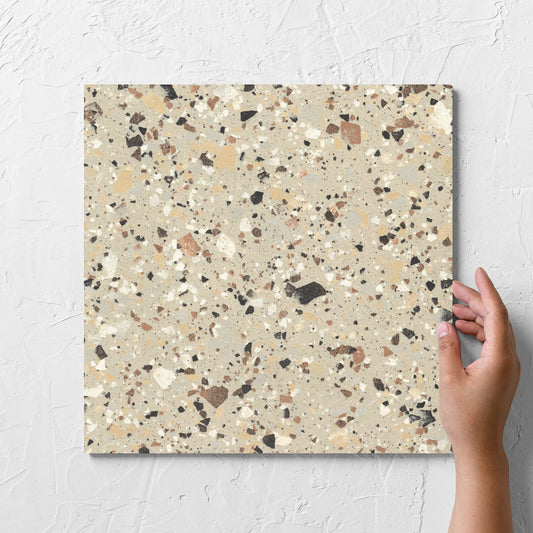 Hand holding a single Venice Beige 300x300mm Matt Terrazzo Tile tile against a white rendered wall, showing the beige matt surface