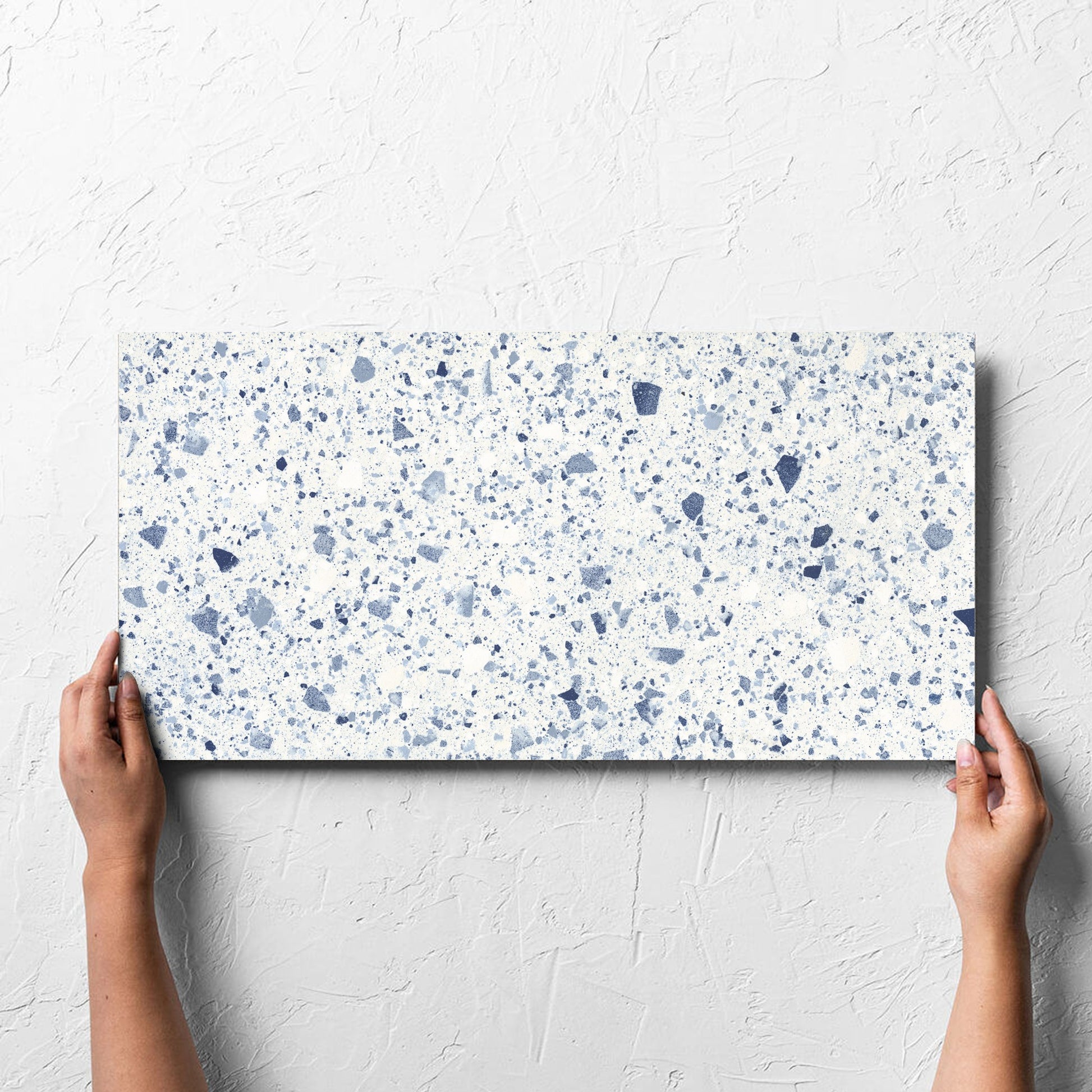 Hand holding a single Venice Blue 300x600mm Matt Terrazzo Tile against a white rendered wall, showing the blue matt surface
