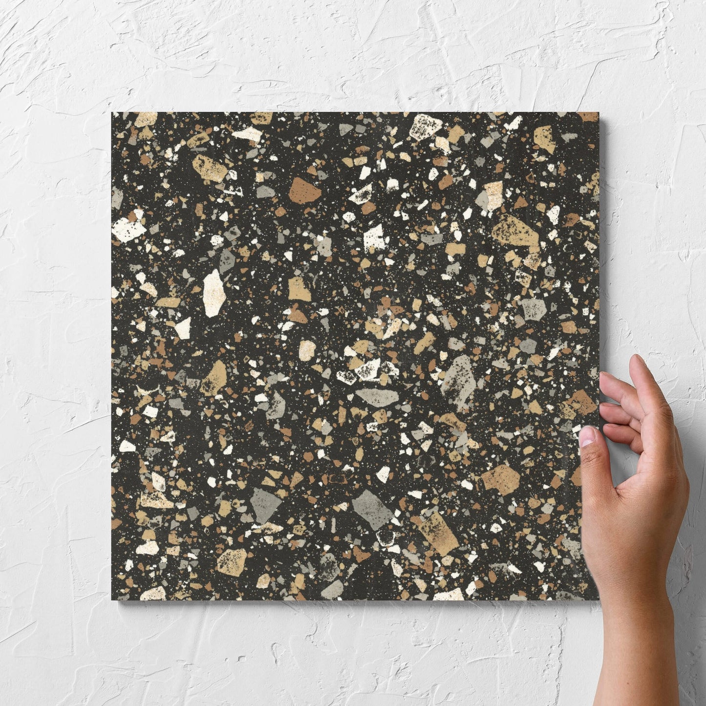 Hand holding a single Venice Earth 300x300mm Matt Terrazzo Tile against a white rendered wall, showing the earth matt surface