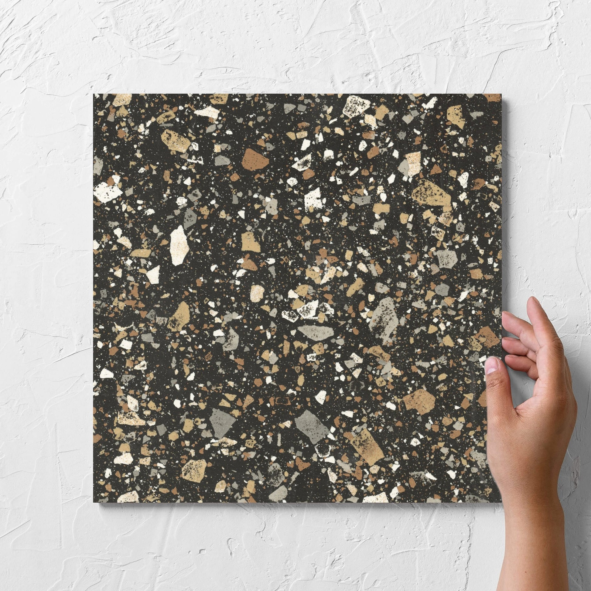 Hand holding a single Venice Earth 300x300mm Matt Terrazzo Tile against a white rendered wall, showing the earth matt surface