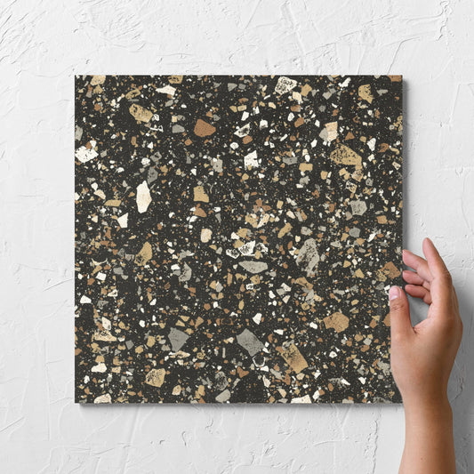 Hand holding a single Venice Earth 300x300mm Matt Terrazzo Tile against a white rendered wall, showing the earth matt surface