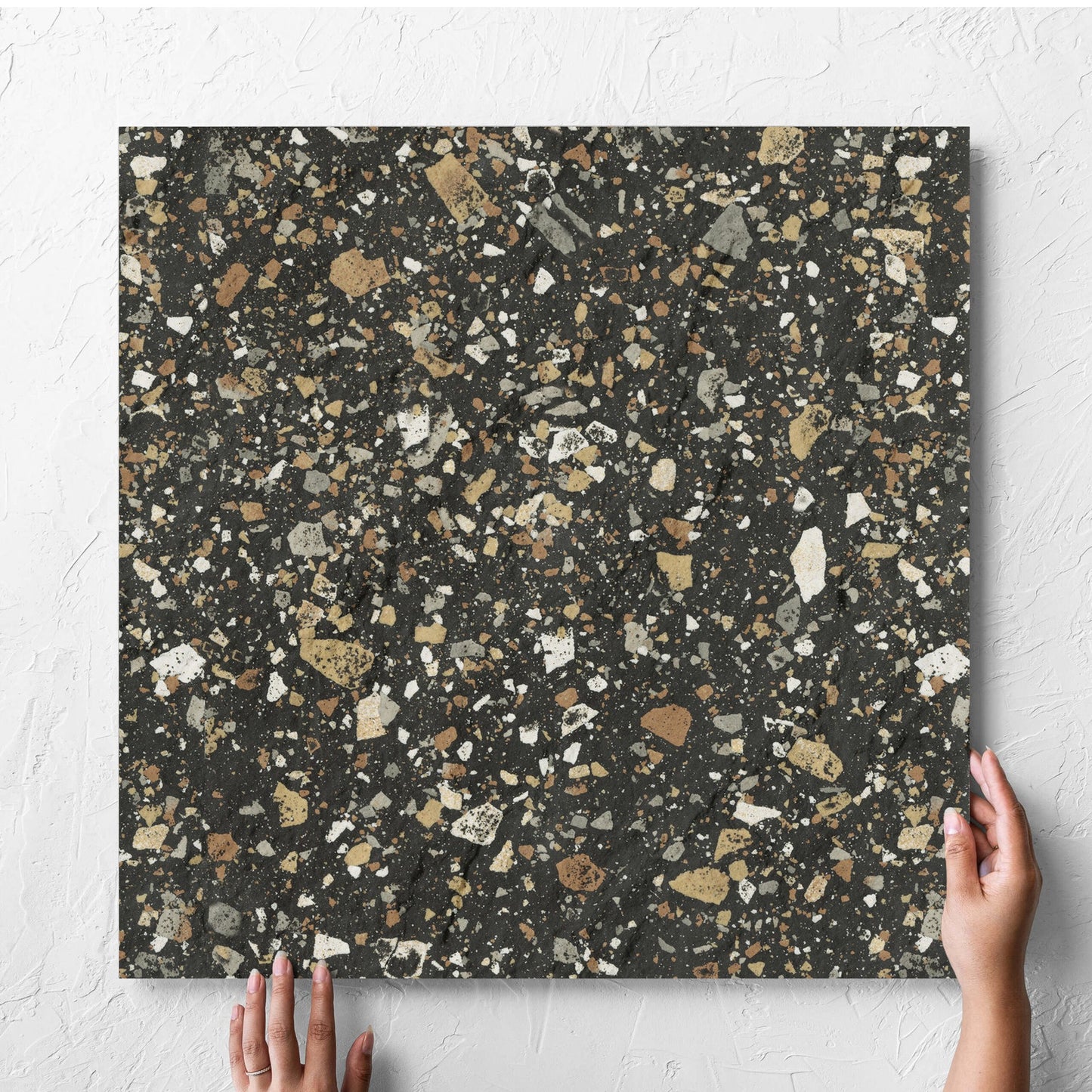 Hand holding a single Venice Earth 600x600mm Matt Terrazzo External Tile against a white rendered wall, showing the earth matt surface