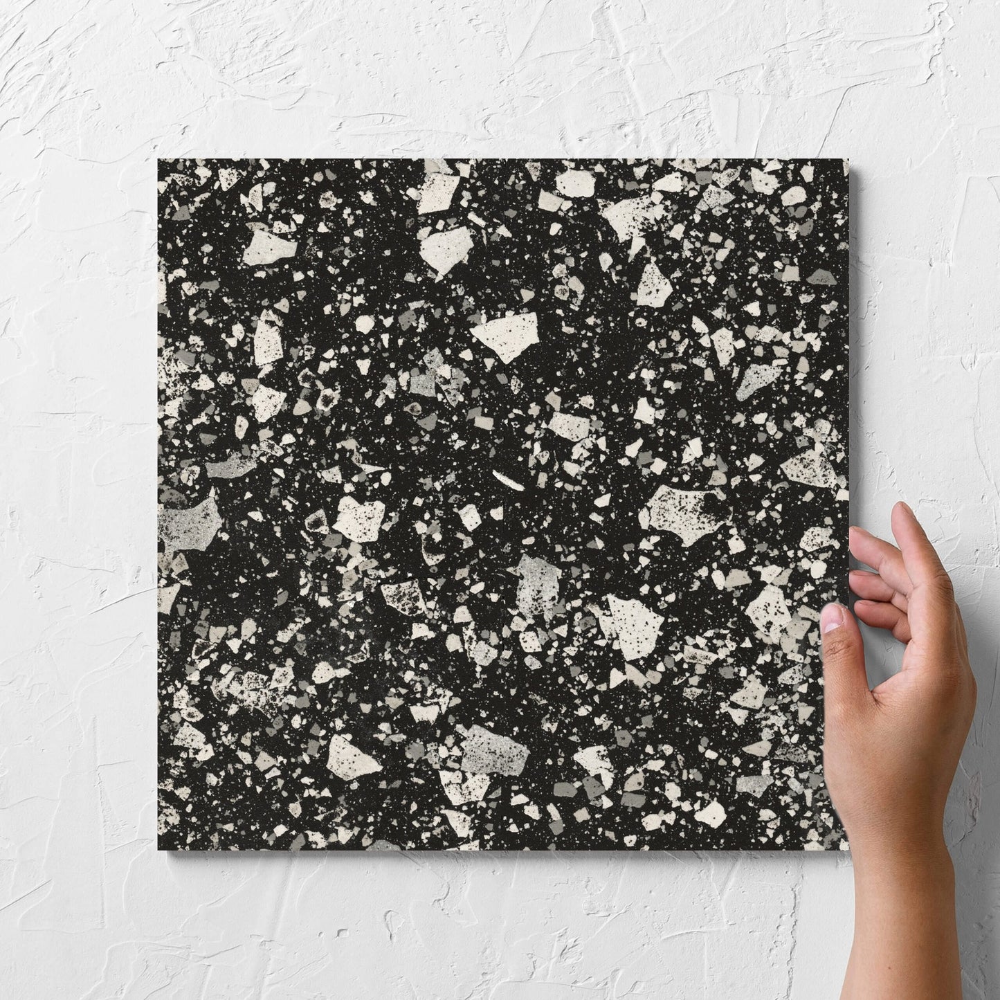 Hand holding a single Venice Graphite 300x300mm Matt Terrazzo Tile against a white rendered wall, showing the graphite matt surface