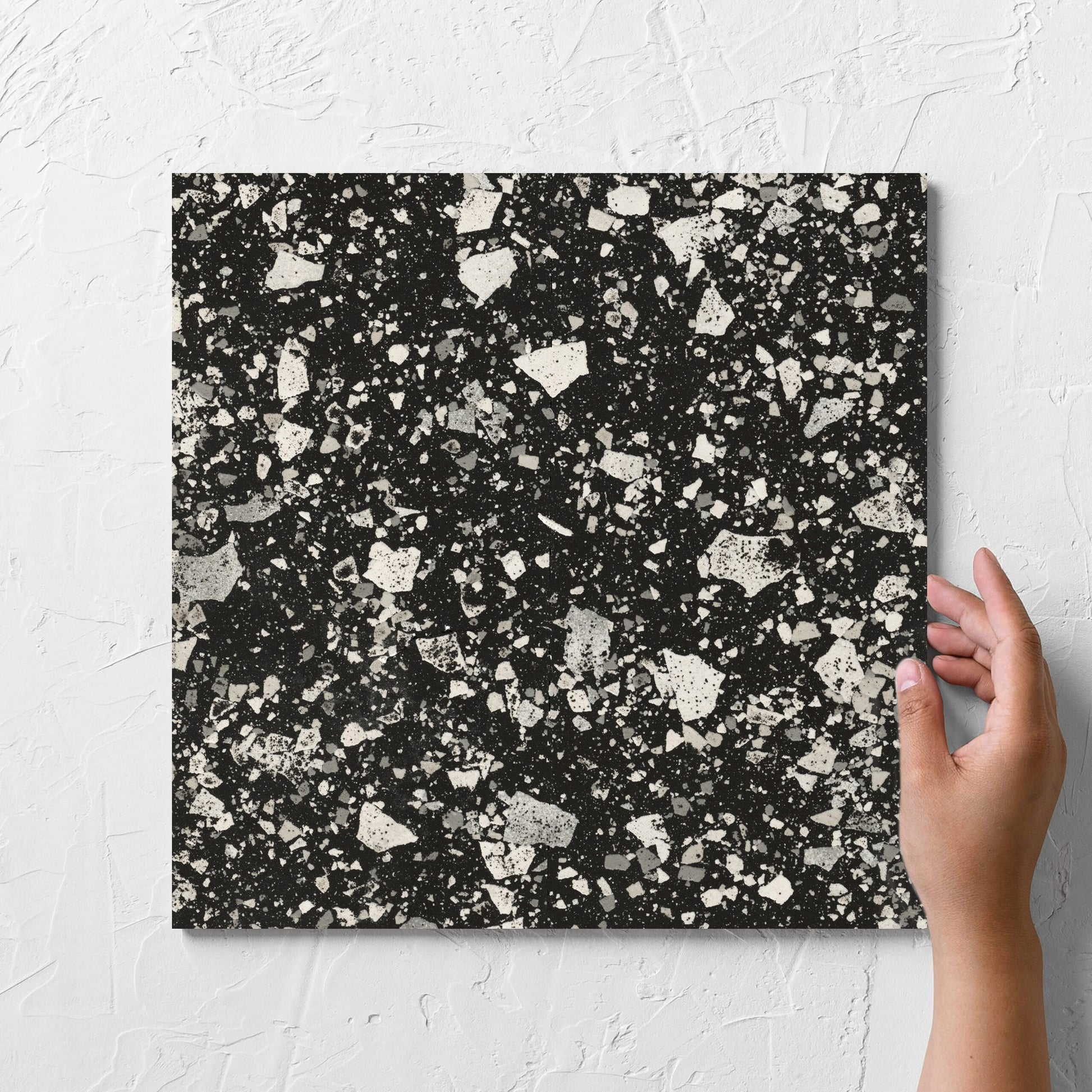 Hand holding a single Venice Graphite 300x300mm Matt Terrazzo Tile against a white rendered wall, showing the graphite matt surface