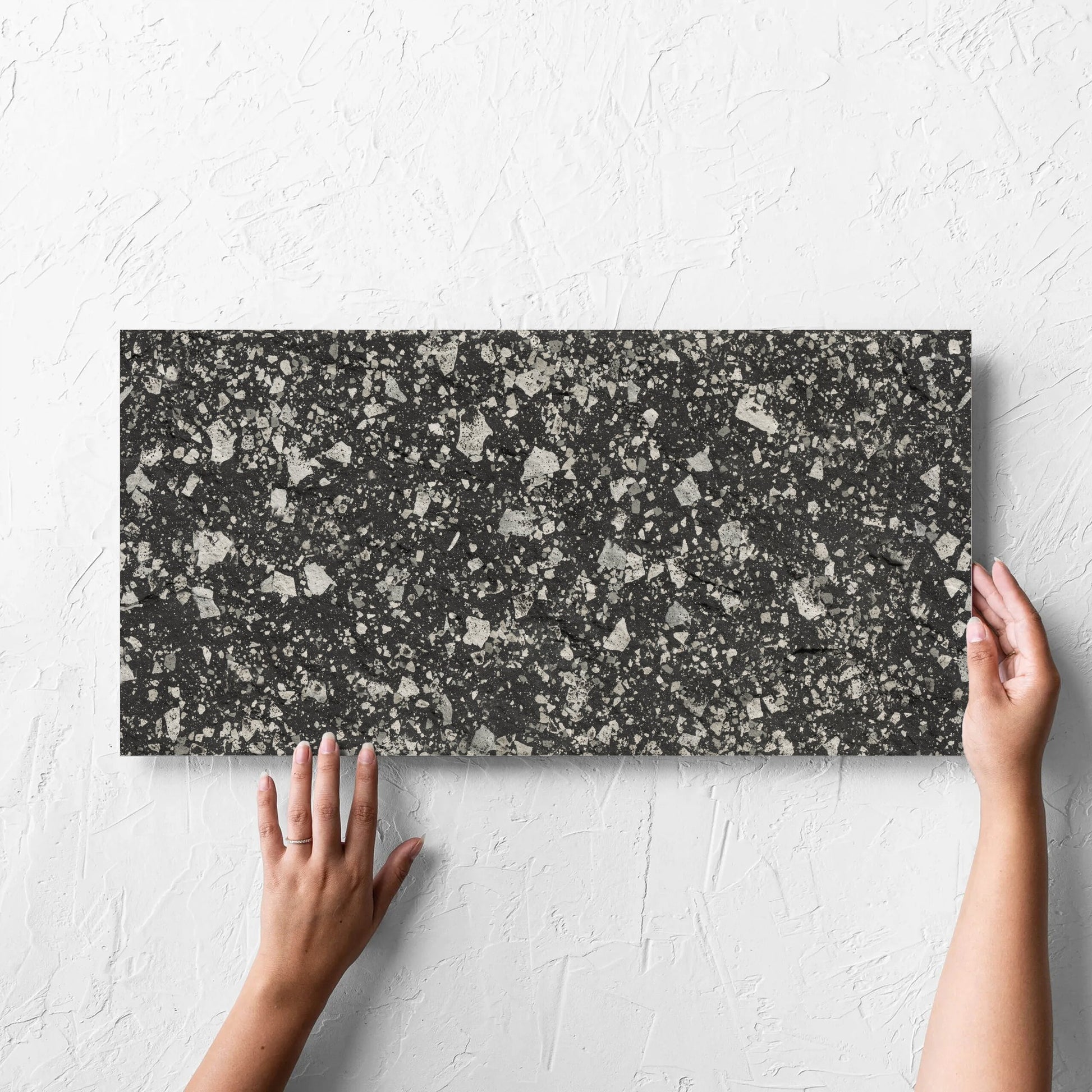 Hand holding a single Venice Graphite 300x600mm Matt Terrazzo External Tile against a white rendered wall, showing the graphite matt surface