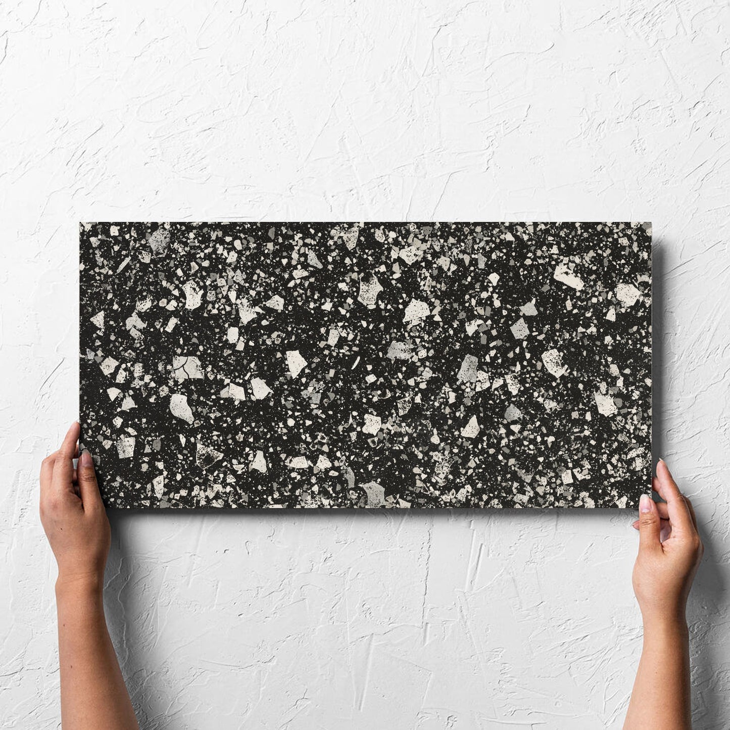 Hand holding a single Venice Graphite 300x600mm Matt Terrazzo Tile against a white rendered wall, showing the graphite matt surface