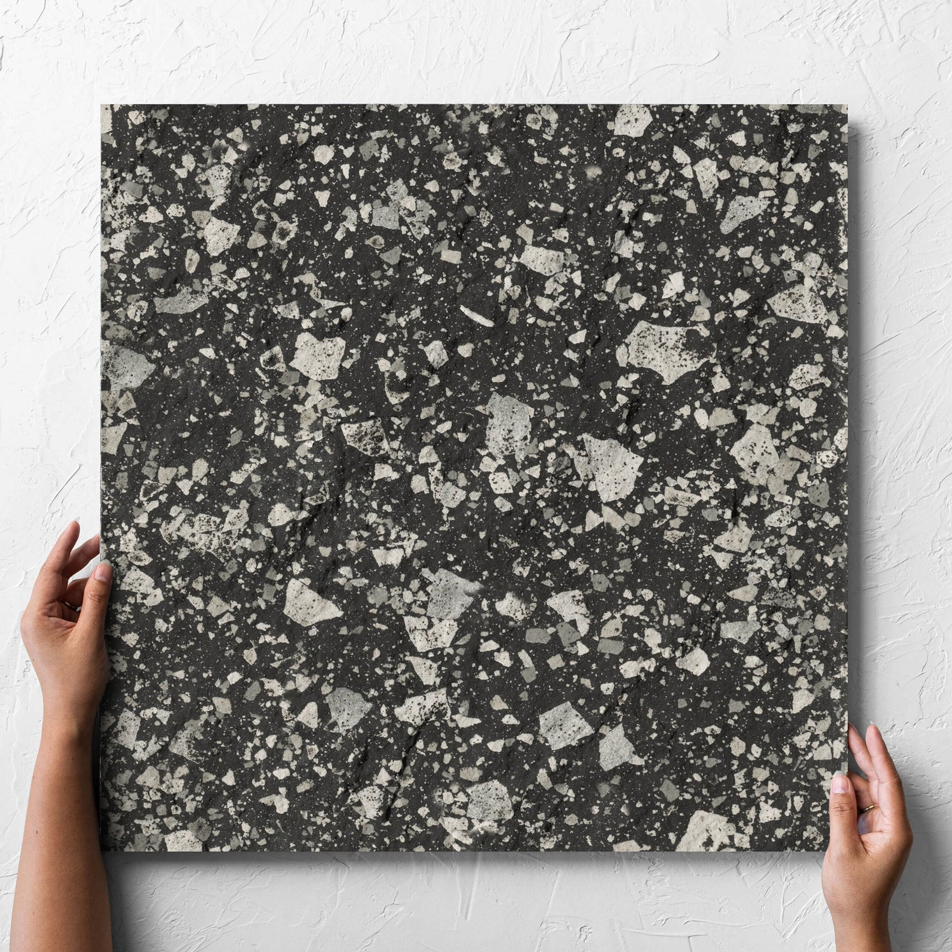 Hand holding a single Venice Graphite 600x600mm Matt Terrazzo External Tile against a white rendered wall, showing the graphite matt surface