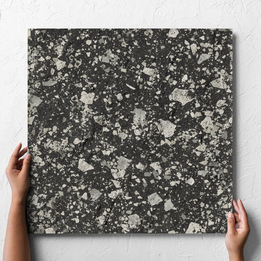 Hand holding a single Venice Graphite 600x600mm Matt Terrazzo External Tile against a white rendered wall, showing the graphite matt surface