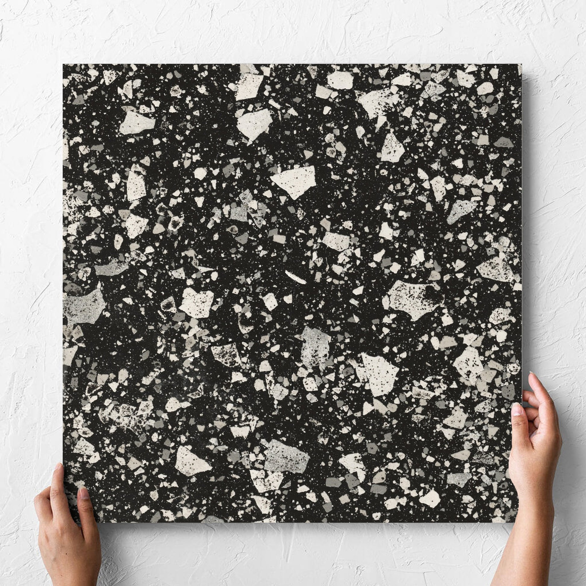 Hand holding a single Venice Graphite 600x600mm Matt Terrazzo Tile against a white rendered wall, showing the graphite matt surface