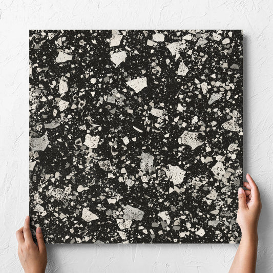 Hand holding a single Venice Graphite 600x600mm Matt Terrazzo Tile against a white rendered wall, showing the graphite matt surface