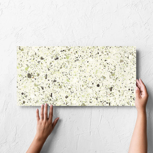 Hand holding a single Venice Green 300x600mm Matt Terrazzo Tile against a white rendered wall, showing the green matt surface