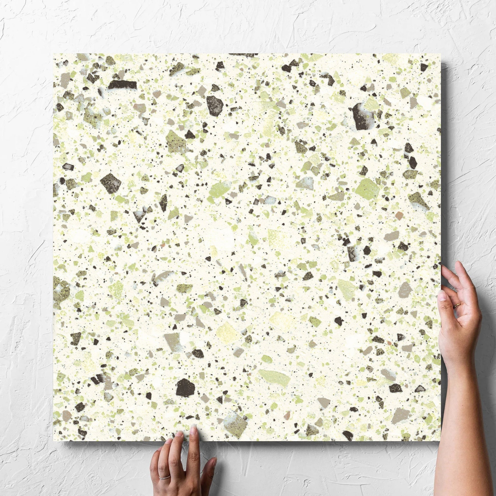 Hand holding a single Venice Green 600x600mm Matt Terrazzo Tile against a white rendered wall, showing the green matt surface