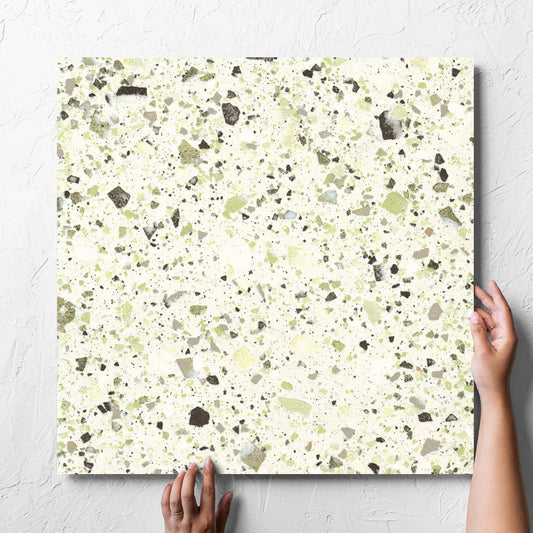 Hand holding a single Venice Green 600x600mm Matt Terrazzo Tile against a white rendered wall, showing the green matt surface