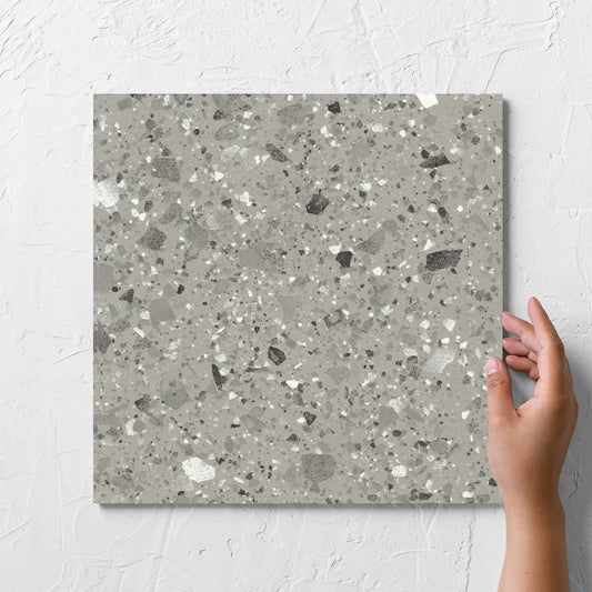 Hand holding a single Venice Grey 300x300mm Matt Terrazzo Tile against a white rendered wall, showing the grey matt surface