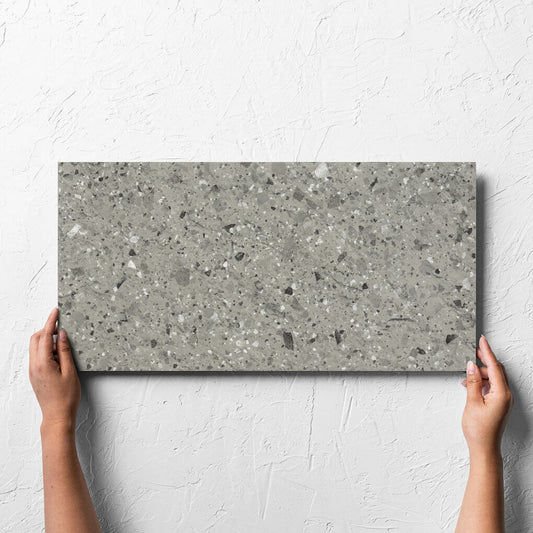 Hand holding a single Venice Grey 300x600mm Matt Terrazzo External Tile against a white rendered wall, showing the grey matt surface