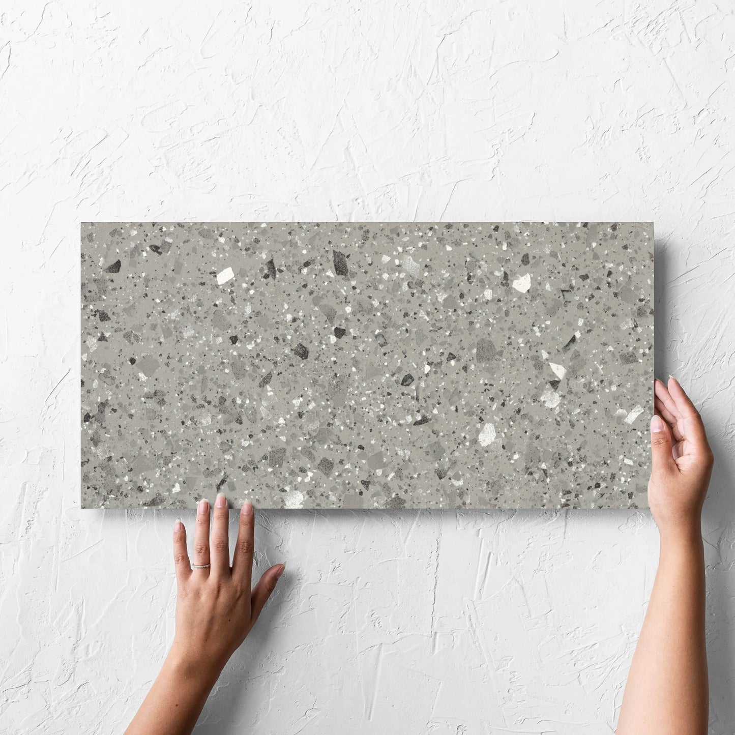 Hand holding a single Venice Grey 300x600mm Matt Terrazzo Tile against a white rendered wall, showing the grey matt surface