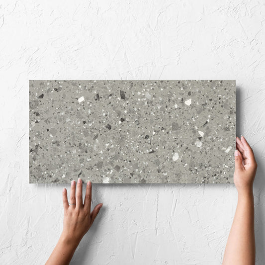 Hand holding a single Venice Grey 300x600mm Matt Terrazzo Tile against a white rendered wall, showing the grey matt surface
