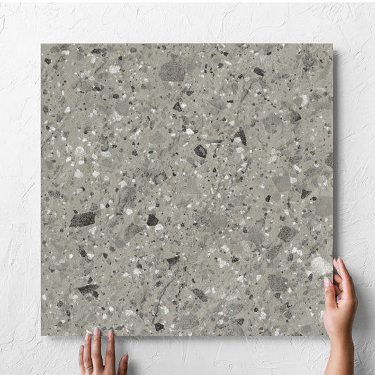 Hand holding a single Venice Grey 600x600mm Matt Terrazzo External Tile against a white rendered wall, showing the grey matt surface
