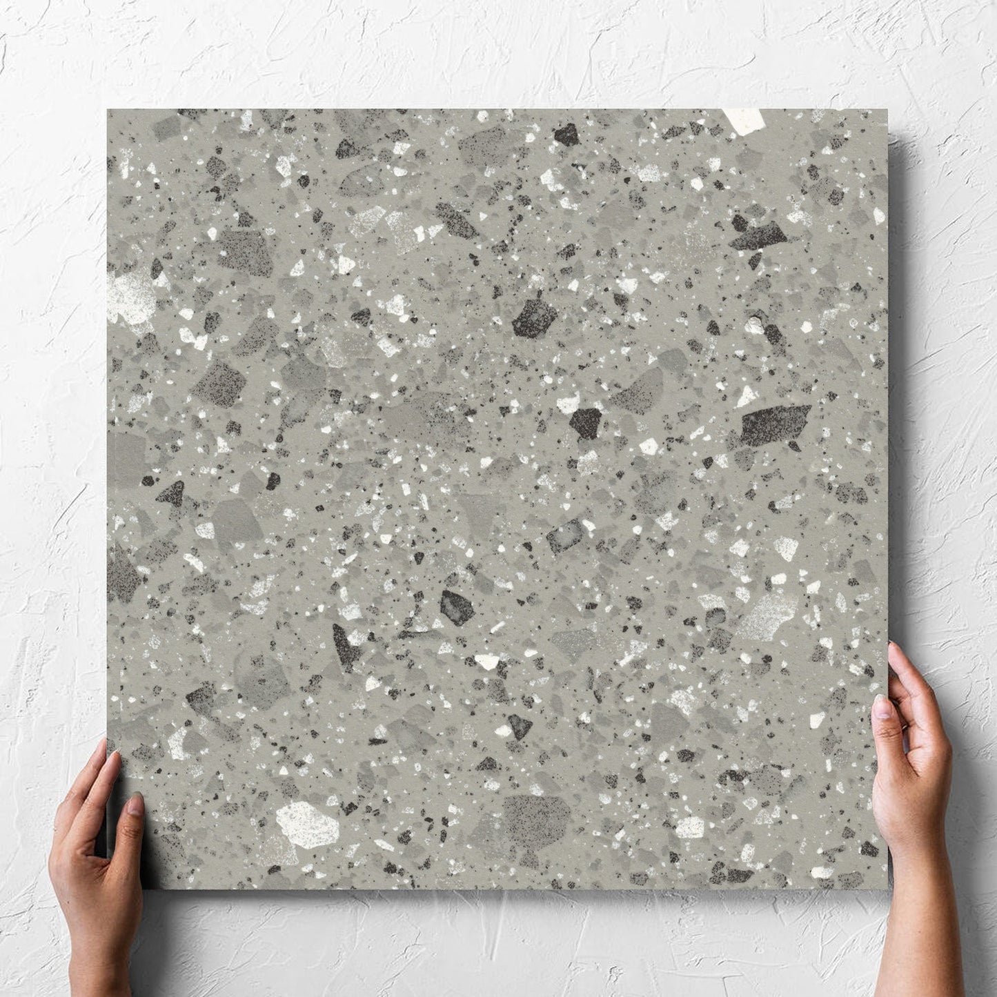Hand holding a single Venice Grey 600x600mm Matt Terrazzo Tile against a white rendered wall, showing the grey matt surface