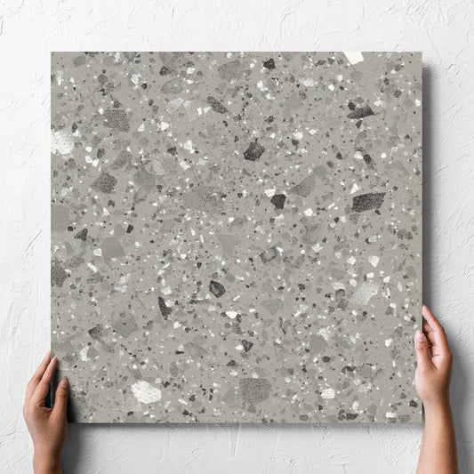 Hand holding a single Venice Grey 600x600mm Matt Terrazzo Tile against a white rendered wall, showing the grey matt surface
