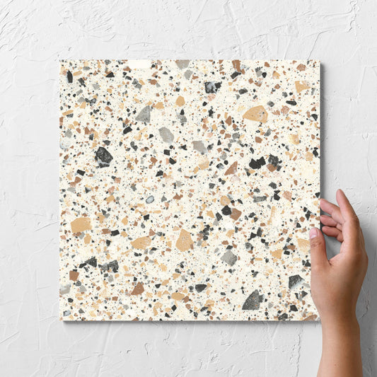 Hand holding a single Venice Ivory 300x300mm Matt Terrazzo Tile against a white rendered wall, showing the ivory matt surface