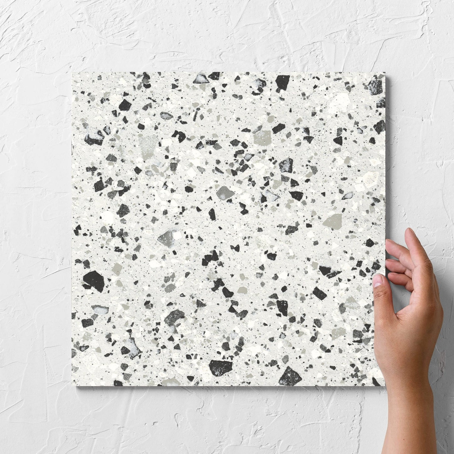 Hand holding a single Venice Silver 300x300mm Matt Terrazzo Tile against a white rendered wall, showing the silver matt surface
