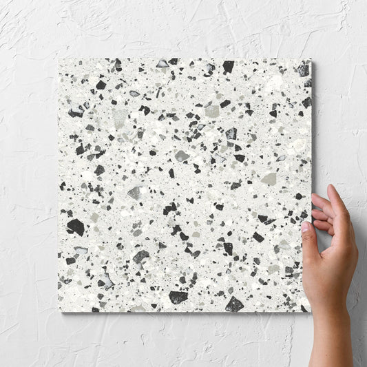 Hand holding a single Venice Silver 300x300mm Matt Terrazzo Tile against a white rendered wall, showing the silver matt surface