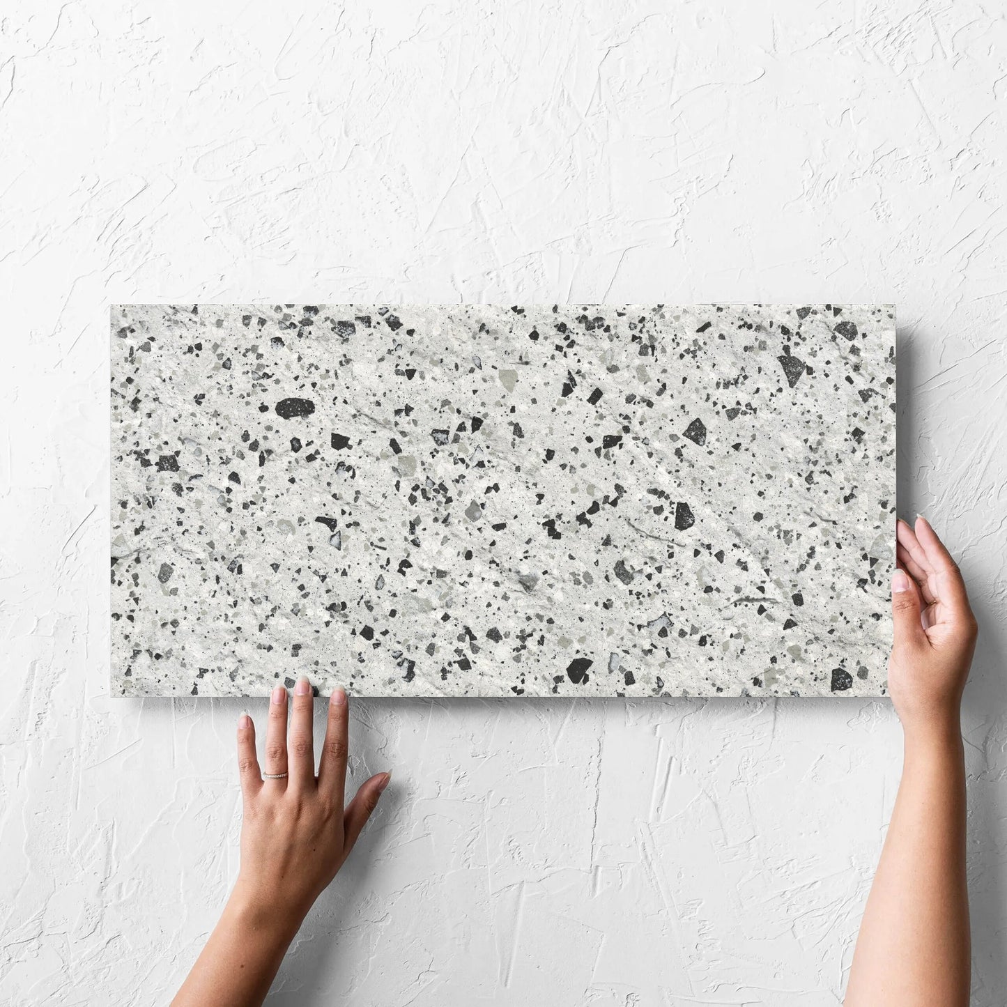 Hand holding a single Venice Silver 300x600mm Matt Terrazzo External Tile against a white rendered wall, showing the silver matt surface