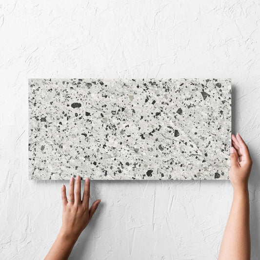 Hand holding a single Venice Silver 300x600mm Matt Terrazzo External Tile against a white rendered wall, showing the silver matt surface