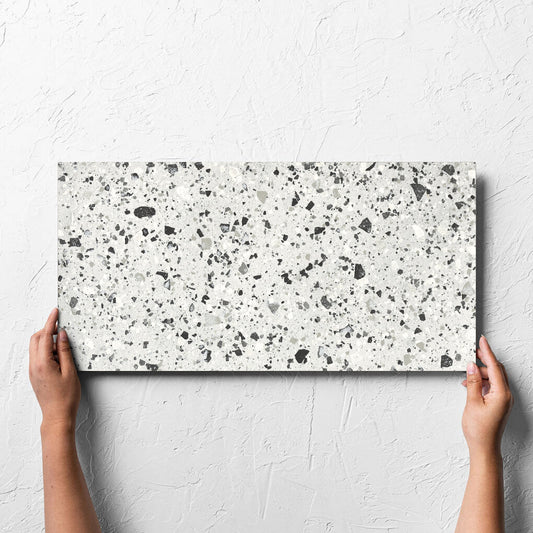 Hand holding a single Venice Silver 300x600mm Matt Terrazzo Tile against a white rendered wall, showing the silver matt surface
