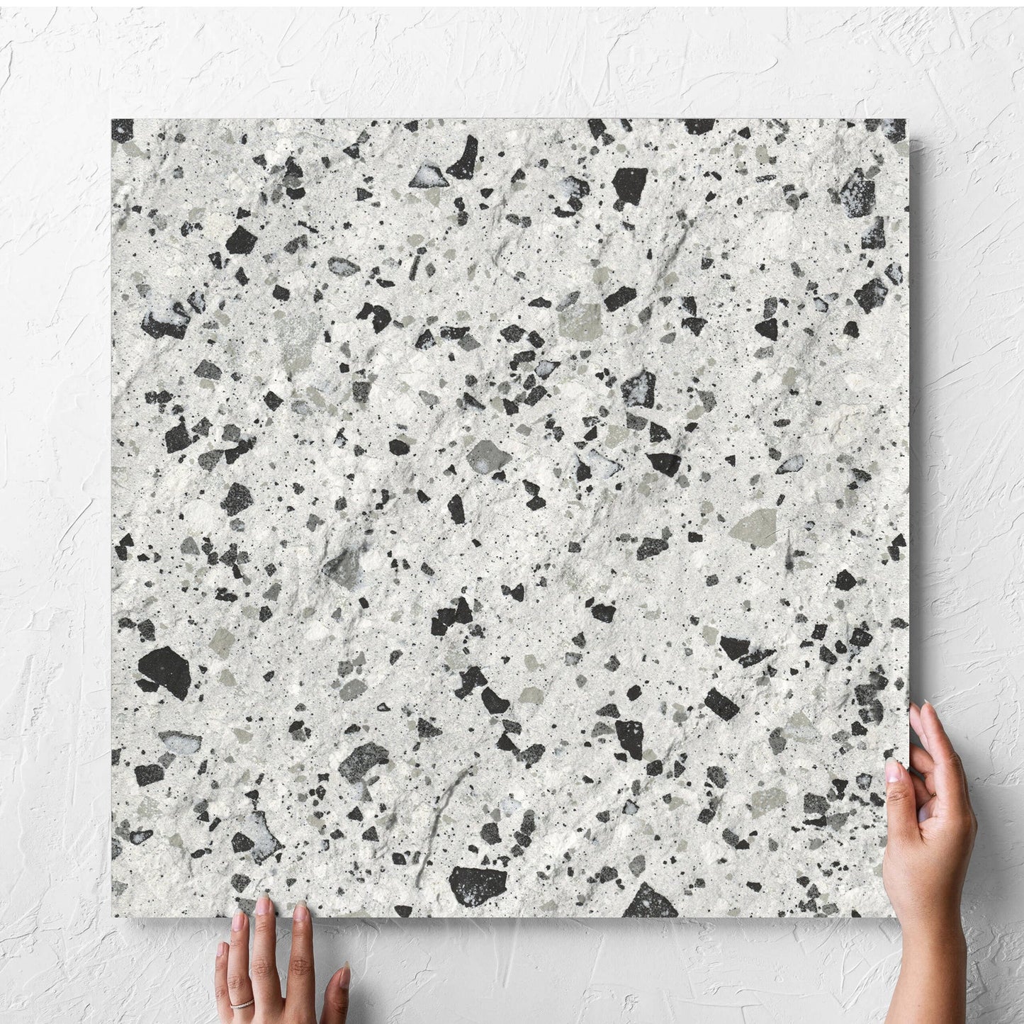 Hand holding a single Venice Silver 600x600mm Matt Terrazzo External Tile against a white rendered wall, showing the silver matt surface