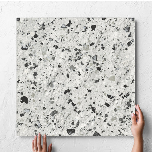 Hand holding a single Venice Silver 600x600mm Matt Terrazzo External Tile against a white rendered wall, showing the silver matt surface