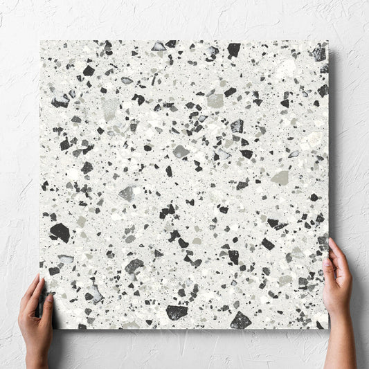 Hand holding a single Venice Silver 600x600mm Matt Terrazzo Tile against a white rendered wall, showing the silver matt surface