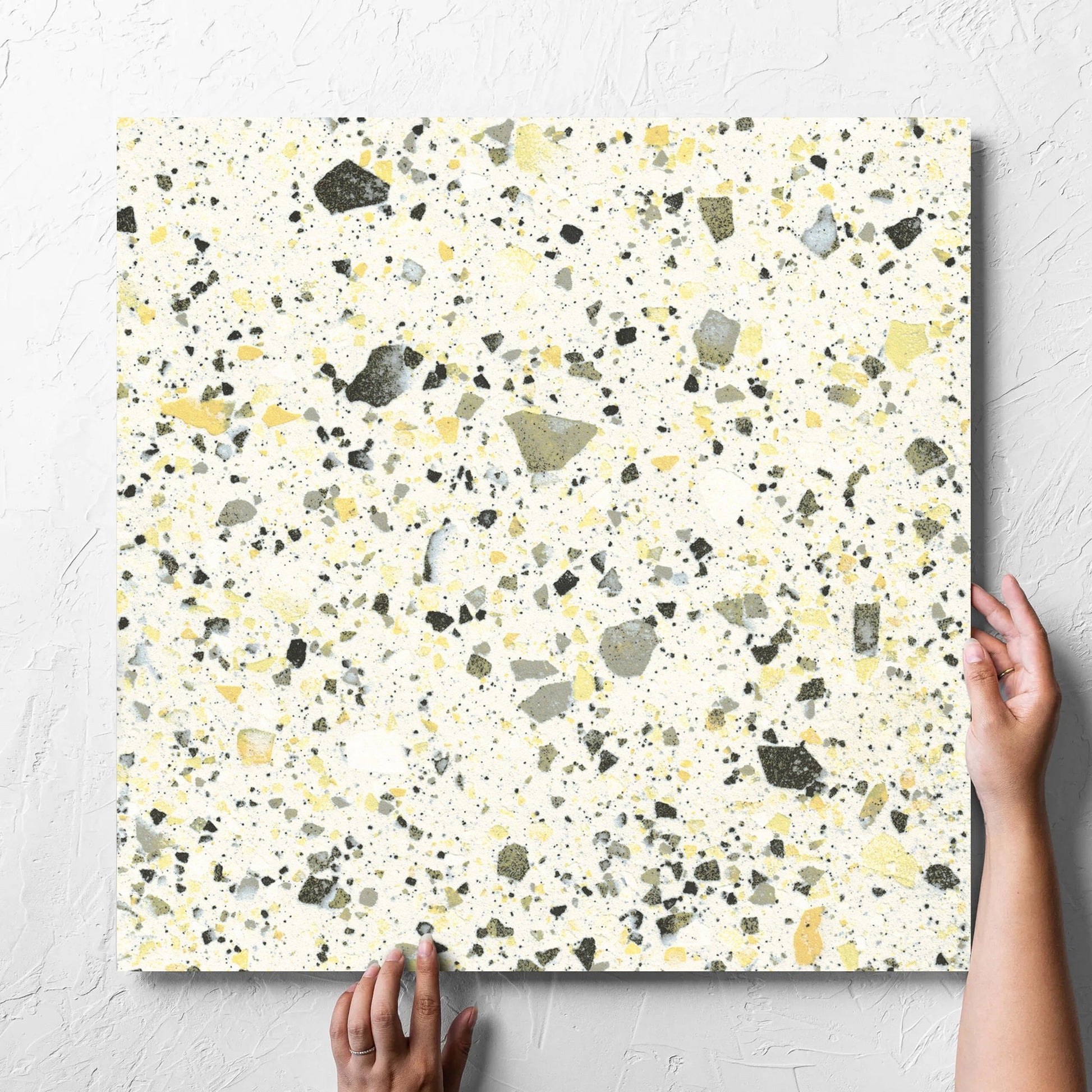 Hand holding a single Venice Yellow 600x600mm Matt Terrazzo Tile against a white rendered wall, showing the yellow matt surface